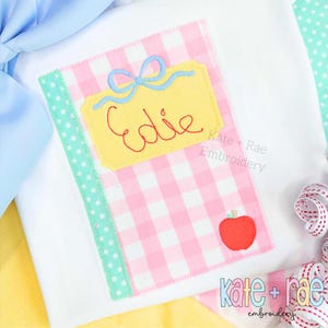 May include: White t-shirt with an embroidered design of a notebook. The notebook has a pink and white gingham pattern, a yellow label with the name "Edie" in red, a blue bow, and a red apple. The design includes the text "Kate + Rae Embroidery".