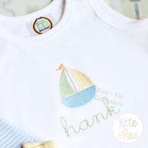 May include: White baby onesie with an embroidered sailboat design in blue, green, and yellow. The name "hank" is embroidered in green cursive below the sailboat. The onesie has a tag that says "Baska Boutique".