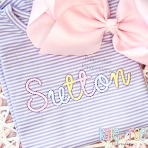 May include: A light purple and white striped shirt with the name "Sutton" embroidered in pastel colors. A large pink bow is at the top. The text "Kate + Rae Embroidery" is also visible.