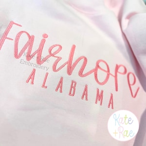 May include: A light pink sweatshirt with the embroidered word "Fairhope" in a cursive font, with "Alabama" below in block letters. The text is in a coral color. The brand name "Kate + Rae" is also embroidered.