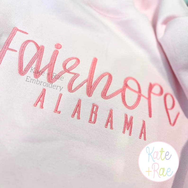 May include: A light pink sweatshirt with the embroidered word "Fairhope" in a cursive font, with "Alabama" below in block letters. The text is in a coral color. The brand name "Kate + Rae" is also embroidered.