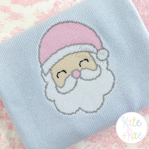 May include: A light blue knitted blanket featuring a central graphic of a Santa Claus face with a pink hat, a beige face, and a white beard. The design includes a small pink nose and closed eyes. A circular logo with the text Kate + Rae is visible.