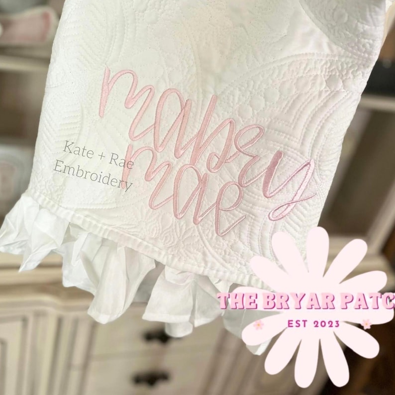 May include: White quilted blanket with pink embroidered text that reads "Maybe Mae." The blanket has a ruffled edge and the text "Kate + Rae Embroidery" is also visible. The logo for "The Bryar Patch" is in the bottom right corner.