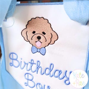 May include: A white fabric bib with light blue trim and straps featuring an embroidered tan poodle wearing a light blue bow tie. Below the dog, the text Birthday Boy is embroidered in light blue script. A small Kate + Rae logo is visible.