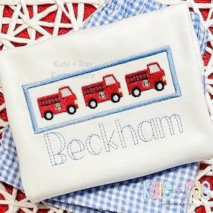 May include: White embroidered baby onesie with a blue border and three red fire trucks. The onesie has the name "Beckham" embroidered in blue below the fire trucks.