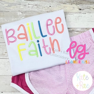 May include: A white t-shirt featuring the embroidered name Baillee Faith in colorful, rounded letters, paired with light pink and white gingham shorts that have a solid dark pink trim along the bottom edges and a white elastic waistband.