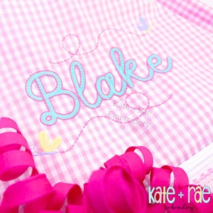 Könnte beinhalten: Rosa-weißes Vichy-Gewebe mit dem Namen "Blake" in hellblauer Schrift gestickt. Die Stickerei hat eine hellblaue Kontur und einen hellrosa Faden. Die Worte "Kate + Rae Embroidery" sind ebenfalls gestickt.