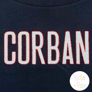 May include: Navy blue fabric with the name "CORBAN" embroidered in white with a red outline. The lower right corner features a circular logo with the text "Kate + Rae" in pastel colors.
