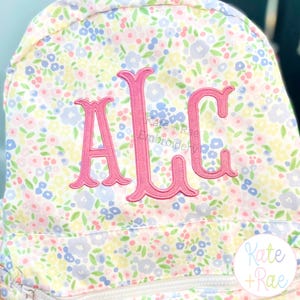 May include: A floral print backpack with a pink embroidered monogram. The backpack features a pattern of small flowers in pastel colours. The monogram includes the letters "ALC". The brand name "Kate + Rae" is also visible.