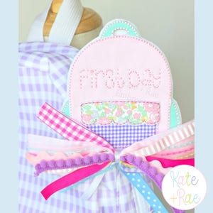 May include: A light pink fabric backpack tag featuring a stitched cupcake design with floral and purple gingham patterns. The tag is adorned with a layered bow made of various ribbons in pink, purple, and blue, including pom-pom and gingham trim.