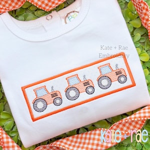 May include: A white onesie with an orange embroidered design of three tractors. The text "Kate + Rae Embroidery" is embroidered on the onesie.