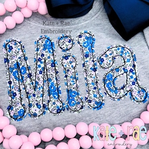 May include: A gray sweatshirt with the name "Mila" embroidered in a floral pattern of blue and white flowers. The text "Kate + Rae Embroidery" is above and below the name. Pink beads and a navy blue bow are also visible.
