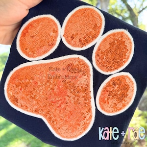 May include: Orange and white paw print design on a navy blue fabric. The paw print is made of sequins and has the text "Kate + Rae Embroidery" in white.