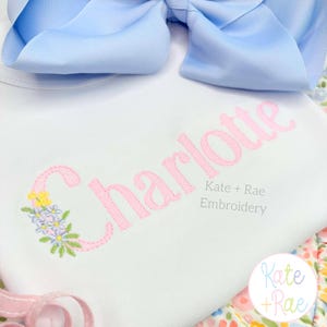 May include: White onesie with the name "Charlotte" embroidered in pink script, accented with a floral design. A large light blue bow sits above the onesie. The text "Kate + Rae Embroidery" is below the name.