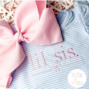 May include: A light blue and white striped baby onesie with the embroidered text "lil sis." A large pink bow is in the upper left corner. The onesie has a round neckline and long sleeves.