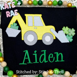 May include: A black t-shirt with a yellow embroidered excavator holding green shamrocks. The name Aiden is embroidered in green below the excavator. The text "Stitched by: Stitch & Thell" is at the bottom of the shirt.