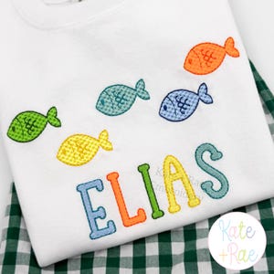May include: White t-shirt with colorful embroidered fish and the name "ELIAS" in blue, orange, green, and yellow. The shirt is folded on a green and white checkered fabric.