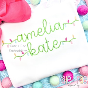 May include: White long-sleeved shirt with green embroidered text "amelia kate" with a string of colourful lights. The text is from Kate + Rae Embroidery.
