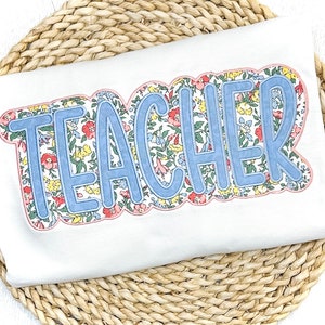 May include: A white t-shirt with the word "TEACHER" in blue outlined letters. The letters are filled with a floral pattern in shades of pink, yellow, and green.