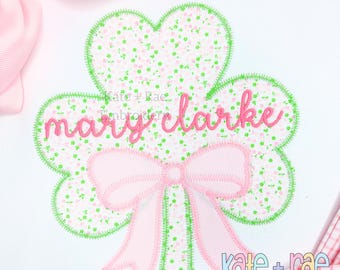 May include: Pink and green embroidered shamrock with a pink bow and the name "mary clarke" in pink lettering.