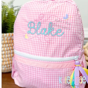 May include: Pink gingham backpack with the name "Blake" embroidered in turquoise script. The backpack features a white zipper, a pink ribbon accent, and a colorful tassel. The text "Kate + Rae Embroidery" is also visible.