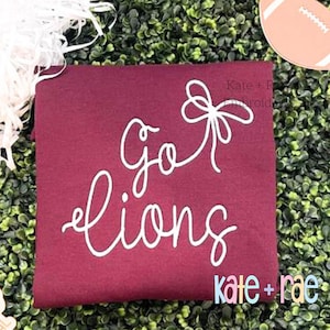 May include: Burgundy t-shirt with white cursive text that reads "go lions" and a white bow. The text "kate + rae" is also visible in the bottom right corner.