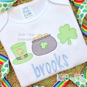 May include: White bodysuit with a green shamrock, a pot of gold, a green leprechaun hat, and the name "Brooks" embroidered in blue. The text "Kate + Rae Embroidery" is also visible.
