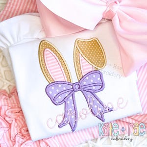 May include: A white shirt with a pink and brown bunny ear design and a purple bow. The name "Caroline" is embroidered in a cursive font below the bunny ears. The shirt is from Kate + Rae Embroidery.