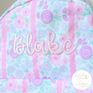 Puede incluir: Un art&iacute;culo de tela con un patr&oacute;n floral repetitivo en tonos azul claro, rosa y lavanda, acentuado por rayas verticales rosas festoneadas. El nombre Blake est&aacute; bordado en el centro en una letra cursiva rosa claro con un contorno azul claro.