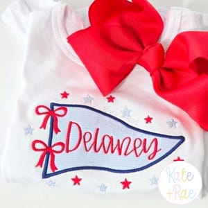 May include: A white short-sleeved shirt featuring an embroidered light blue pennant with a dark blue border. The name Delaney is stitched in red script inside the pennant, accented by red bows and small red and light blue stars. A large red hair bow is attached.