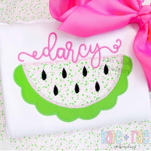 Puede incluir: Una camiseta blanca con una aplicación de sandía rosa y verde y el nombre "darcy" bordado en rosa encima. El texto "Kate + Rae Embroidery" también está bordado en la camiseta.