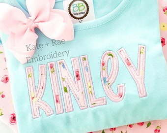 May include: A light blue t-shirt with the name "KINLEY" embroidered in a floral pattern. The letters are outlined in pink. A pink bow is placed on the shirt. The text "Kinley Satin Stitch Applique Font" is at the top and bottom of the image.
