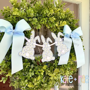 Könnte beinhalten: Ein grüner Kranz mit hellblauen Schleifen und einem Banner mit drei Hasenausschnitten. Die Hasen haben ein Blumenmuster und flauschige weiße Schwänze. Das Banner ist mit einem hellrosa Band verbunden. Der Text "Kate + Rae Embroidery" ist sichtbar.