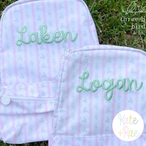 Op de afbeelding: Twee gepersonaliseerde rugzakken met een roze en wit bloemenpatroon en groen geborduurde namen. Op de ene rugzak staat "Laken" en op de andere "Logan". De rugzakken hebben een voorvak met rits en een witte rits. De merknaam "Kate + Rae Embroidery" is zichtbaar.