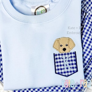Puede incluir: Una camiseta azul claro con un bolsillo de cuadros azules y blancos. La cabeza de un cachorro Labrador amarillo asoma por el bolsillo. El texto "Kate + Rae Embroid" está en la camiseta.