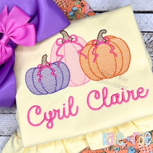 May include: A light yellow shirt embroidered with three pumpkins in purple, pink, and orange, each with a pink bow. The name "Cyril Claire" is embroidered in pink script below the pumpkins. A large purple bow is visible in the background.