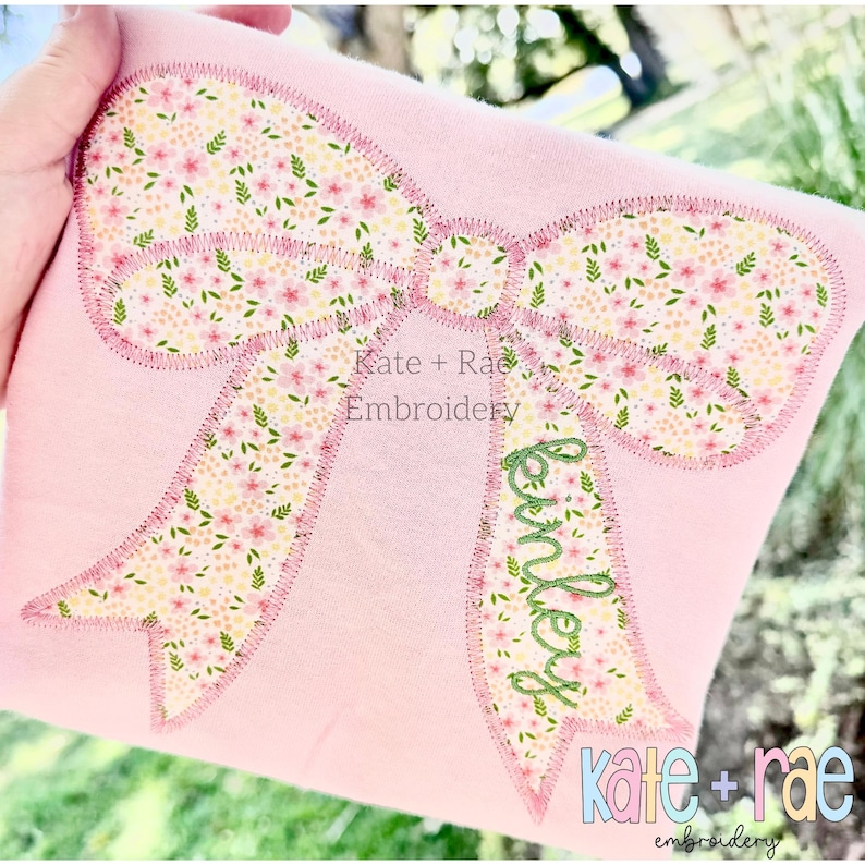Puede incluir: Una camiseta rosa claro con una gran aplicaci&oacute;n de lazo bordado con un estampado floral de peque&ntilde;as flores rosas y hojas verdes. El nombre "Kinley" est&aacute; bordado en cursiva verde en la cinta derecha del lazo.