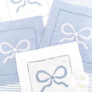 May include: Several square, light blue and white embroidered bow napkins. The napkins have a stitched bow design in the center. The napkins are made of a soft fabric and have a decorative stitched border. The image also shows the text "Kate + Rae Embroidery".