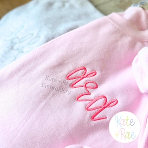 May include: A light pink sweatshirt with the embroidered initials "KW" in a cursive font. The text "Kate + Rae Embroidery" is printed below the initials. A light blue item is visible in the background.