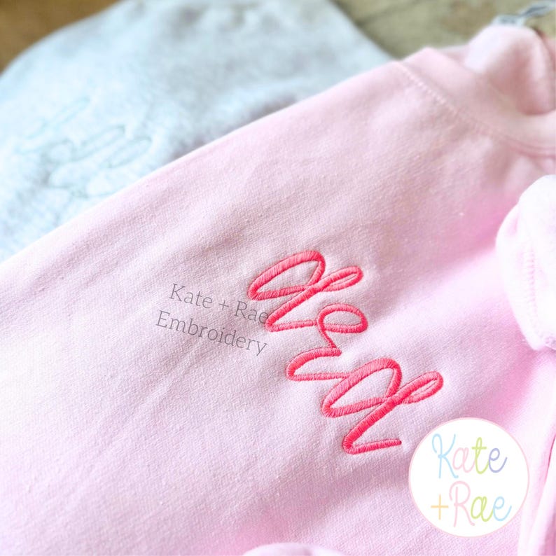 May include: A light pink sweatshirt with the embroidered initials "KW" in a cursive font. The text "Kate + Rae Embroidery" is printed below the initials. A light blue item is visible in the background.