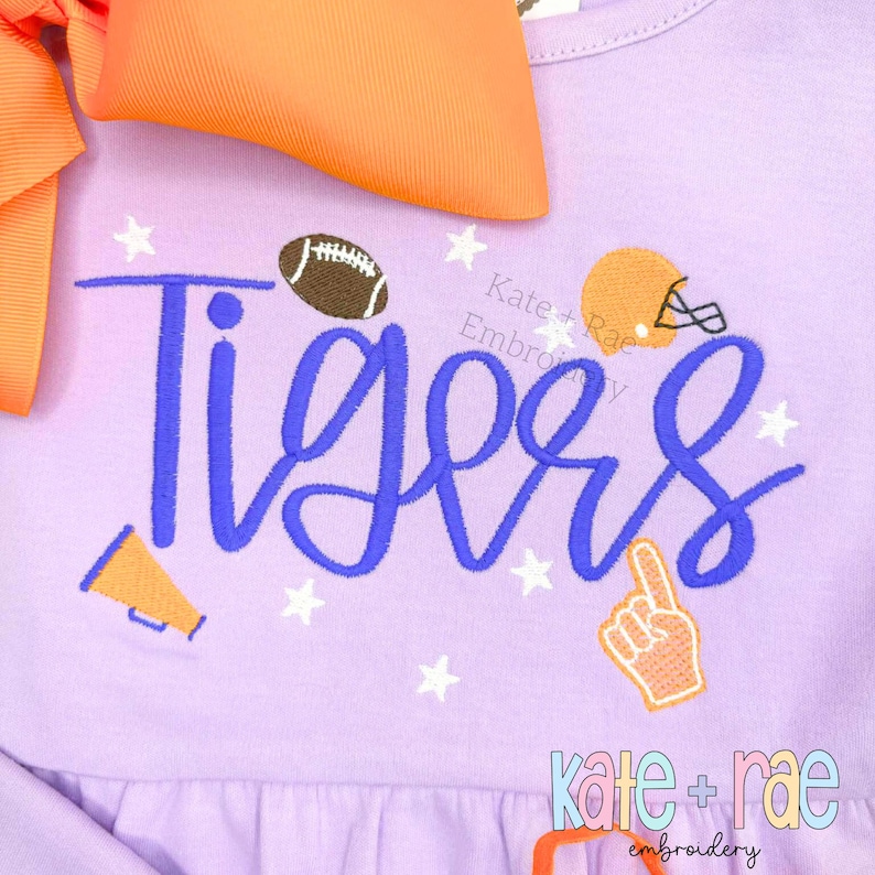 May include: A light purple dress with the word "Tigers" in blue script, featuring a football, helmet, megaphone, and a foam finger. An orange bow is attached to the dress. White stars are scattered around the word "Tigers".