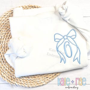 May include: A white long-sleeved shirt with a blue embroidered bow and the number "6" in the centre.