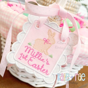 May include: A pink and white embroidered tag with a bunny and the text "Millie's 1st Easter". The tag is attached to a pink ribbon and a white wicker basket.