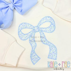 Puede incluir: Una sudadera blanca con un lazo bordado azul con un patrón floral. El texto "kate + rae embroidery" es visible debajo del lazo.