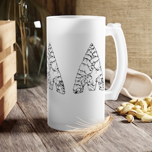 May include: A white frosted glass beer mug with a handle. The mug features two black and white arrowhead designs. The mug is sitting on a wooden surface with a wooden crate, a glass bottle, and pistachios in the background.