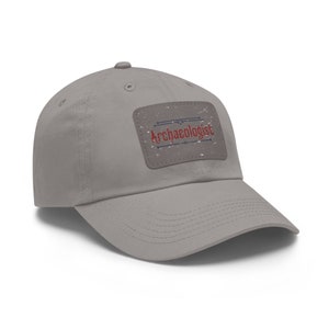 May include: A gray baseball cap with a curved brim. The cap features a rectangular patch with the word "Archaeologist" in red script. The patch has a distressed, vintage look.