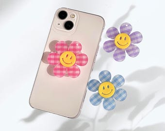 May include: A smartphone with a pink and white checkered flower phone grip attached. Two additional flower grips in purple and blue checkered patterns with yellow smiley faces are also visible.