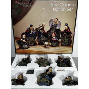 May include: An 8-piece ceramic nativity set in a box labeled Brighten the Season. The set includes figurines of figures in blue and red robes with gold filigree patterns, an angel with wings, and a small manger, all nestled in white foam packaging.