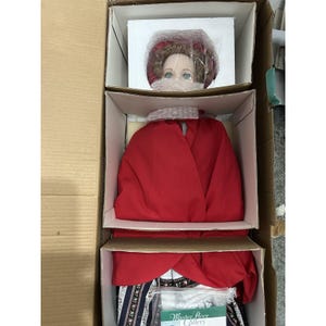 May include: A doll with blue eyes and a red hat, packaged in a cardboard box. The doll's head is visible, wrapped in bubble wrap. The box contains a red garment and a dress with a floral pattern. The text "Master Piece Gallery" is visible.
