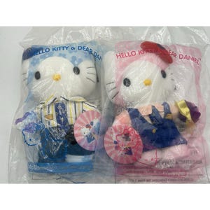 May include: Two Hello Kitty and Dear Daniel plush toys in plastic packaging. One plush is dressed in a striped shirt and blue pants, the other in a pink dress. Each toy has a matching paper fan with a Hello Kitty design.